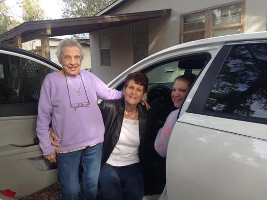 Three family members smile together near a car in front of a house, enjoying their time together.