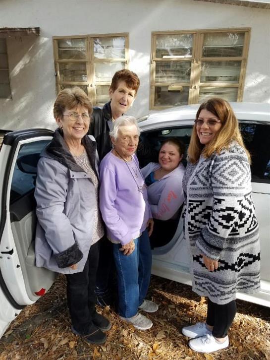 Five women stand together by a white car, sharing smiles and enjoying each other's company outside.