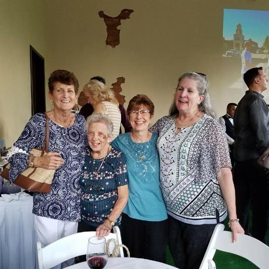 Four women stand together, smiling at a social event, enjoying their time in a lively atmosphere.
