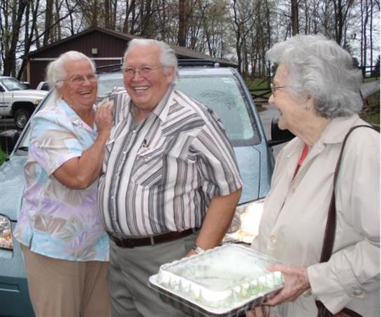 Three elderly friends are smiling and sharing laughter outside a vehicle in nature.