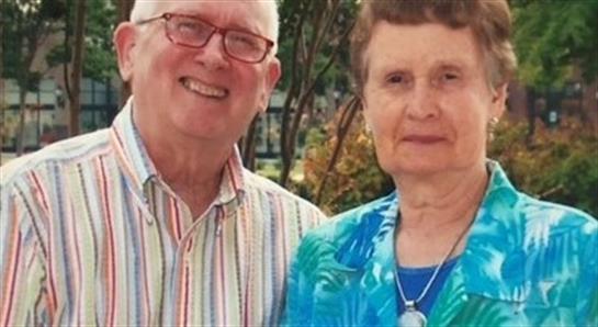 Happy couple stands together in a garden, smiling warmly at the camera on a bright day.