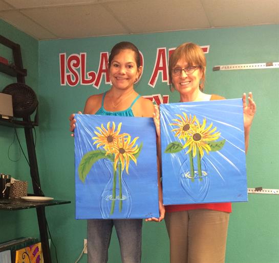 Two women hold up their finished sunflower paintings, excited after a creative art session.