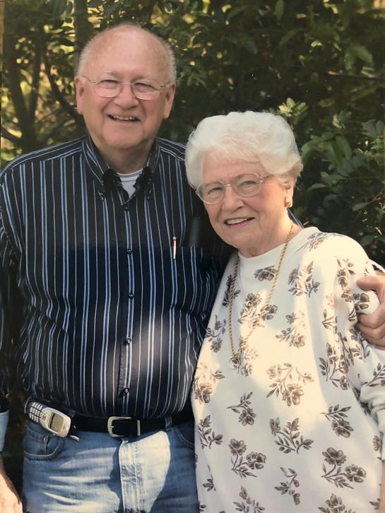 Two smiling seniors stand closely together in a garden with greenery around them.