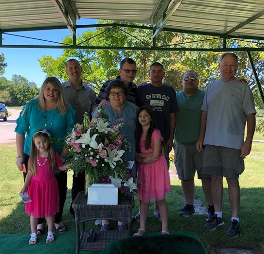 A group of family members gathers in a memorial park under a shelter on a bright day.