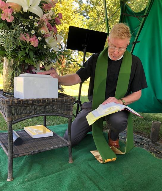 A minister reads from a Bible while kneeling beside a decorative floral display at a ceremony.