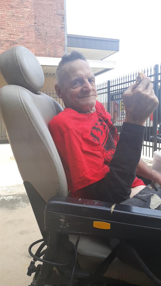 An elderly man smiles and playfully gestures while seated on his mobility scooter outdoors.