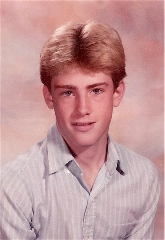 A blonde young man smiles in a classic 1980s school portrait with a soft background.