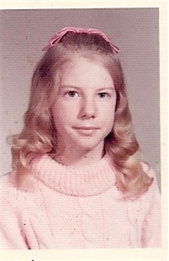 A young girl smiles softly, showcasing her curly hair and a pink sweater in a vintage studio.