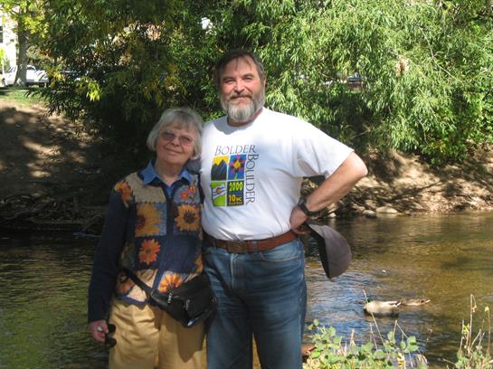 Two people stand together beside a calm stream, enjoying a sunny day surrounded by greenery.