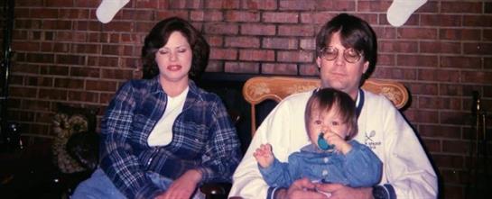 Two adults pose with a baby in a cozy living room filled with warmth and nostalgia.