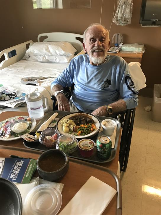 An elderly man sits in a wheelchair at a hospital room table, enjoying a varied meal.