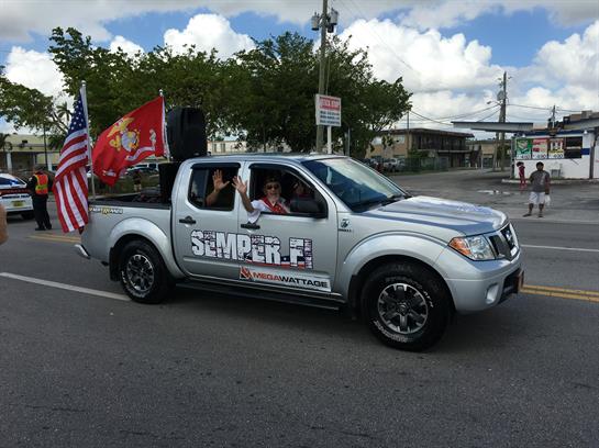 A silver pickup truck adorned with flags is driving through a lively parade with participants.
