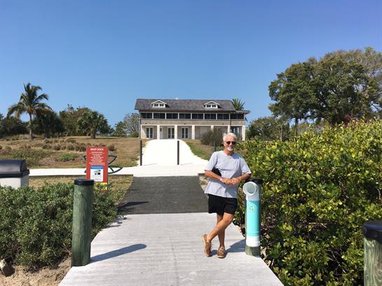 A person stands casually by a pathway that leads to a beach house under clear blue skies.