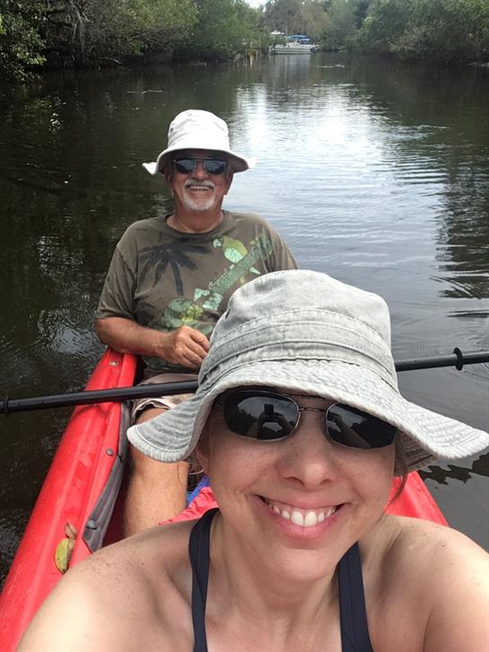 Two people are paddling in a kayak on clear water, smiling under sunny skies while wearing hats.