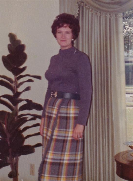 Vintage style captures a woman in a plaid skirt and sweater standing near a large plant indoors.