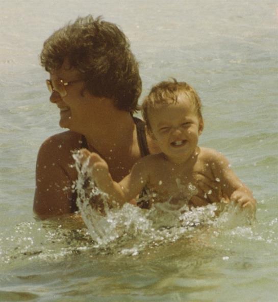 An adult and a child splash joyfully in shallow water during a bright, sunny day at the beach.