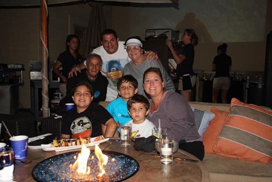 Family gathers around a fire pit, enjoying food, drinks, and laughter in a cozy atmosphere.