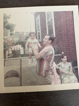 A joyful parent lifts a smiling baby while a woman sits nearby, enjoying a warm afternoon outside.