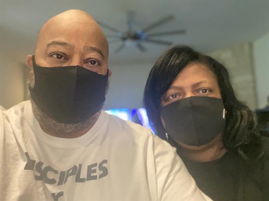 A couple takes a selfie together, both wearing black masks, in their cozy living room.