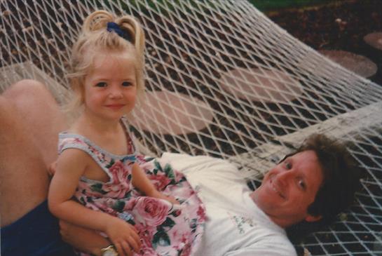 A joyful little girl sits on her father's lap, both relaxing on a hammock in the garden.