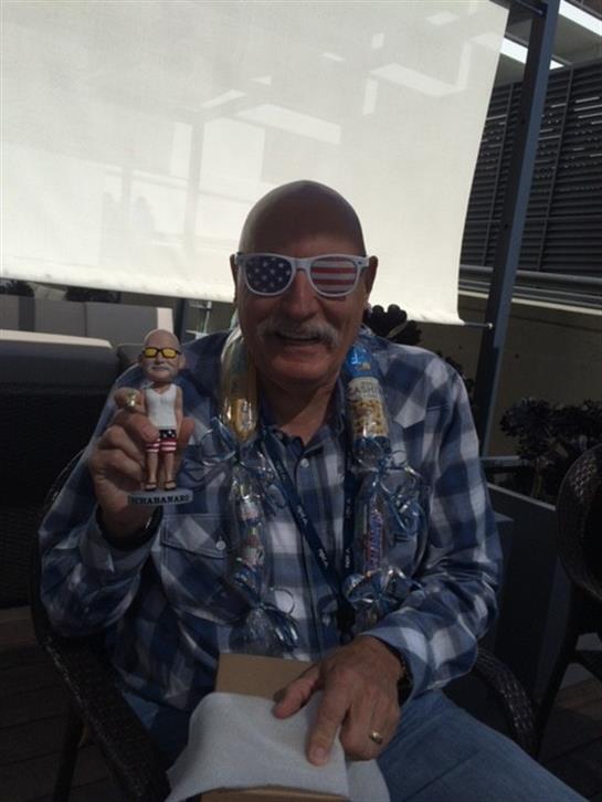 Older man in sunglasses and plaid shirt holds a small figurine, smiling joyfully at the event.