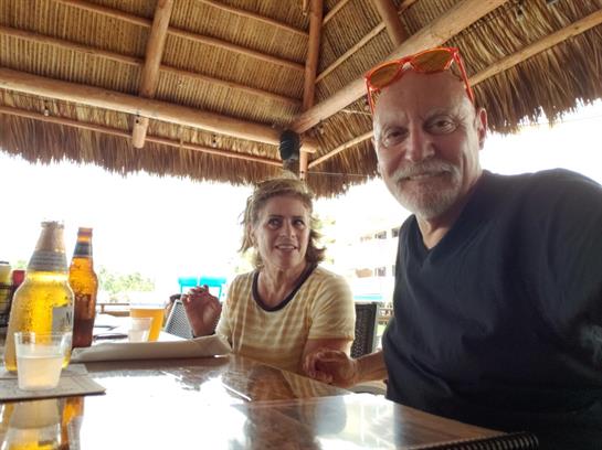 A cheerful couple sits at a table, smiling while enjoying beverages under a thatched roof.