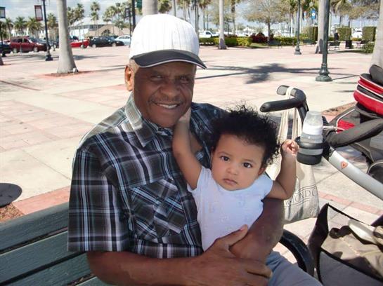 An elderly man smiles while holding a baby in a park, surrounded by palm trees and clear skies.