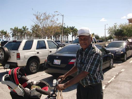 An elderly man stands smiling beside a stroller in a bustling parking lot surrounded by palm trees.