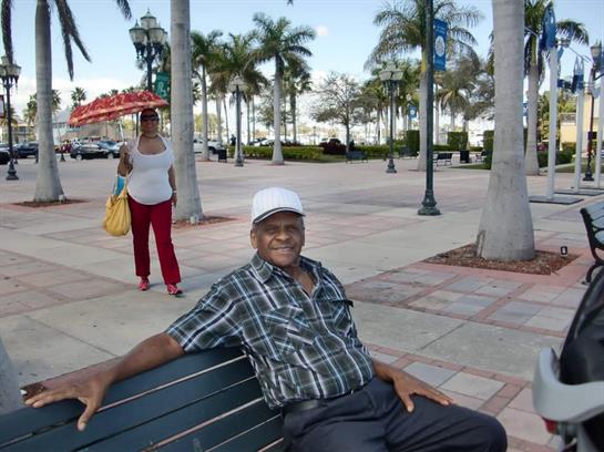 An elderly man relaxes on a park bench with palm trees and a woman walking nearby.