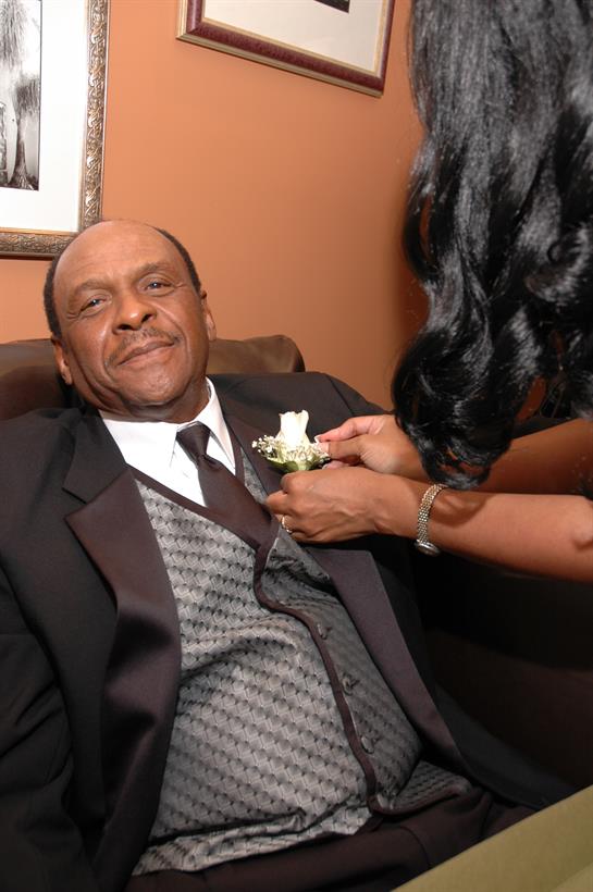 A man in a suit relaxes as a woman pins a flower on his lapel in a cozy setting.