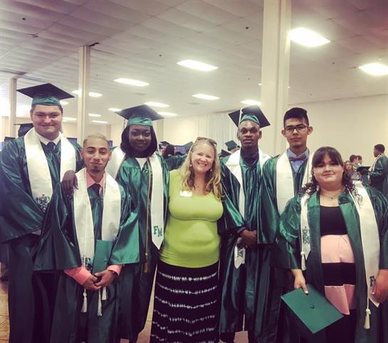 Students in green caps and gowns celebrate graduation, proudly smiling with their teacher.