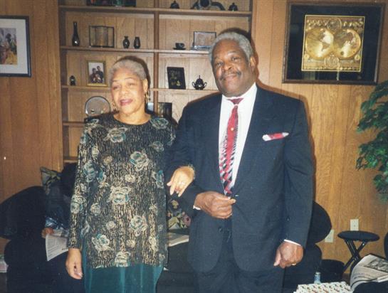 A couple stands side by side in a warmly decorated living room, both smiling and dressed elegantly.