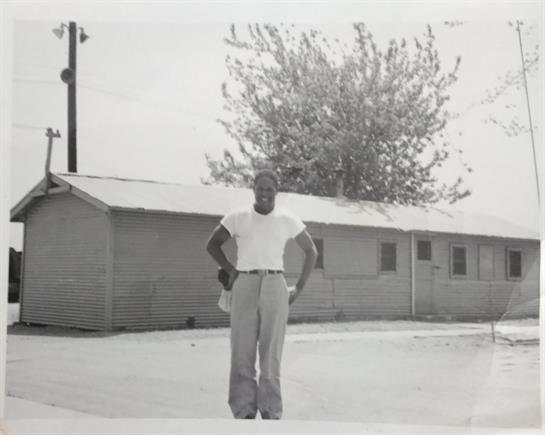 A man is posing proudly in front of a simple building with a sizable tree nearby.