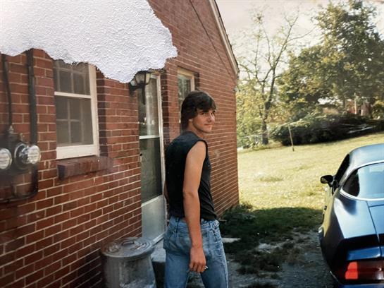 A young person wearing a sleeveless shirt smiles while standing outside a brick home in daylight.