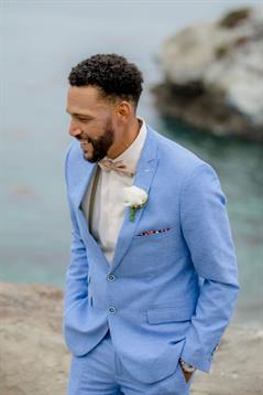 A groom in a light blue suit enjoys the moment at a seaside wedding, smiling as he walks.