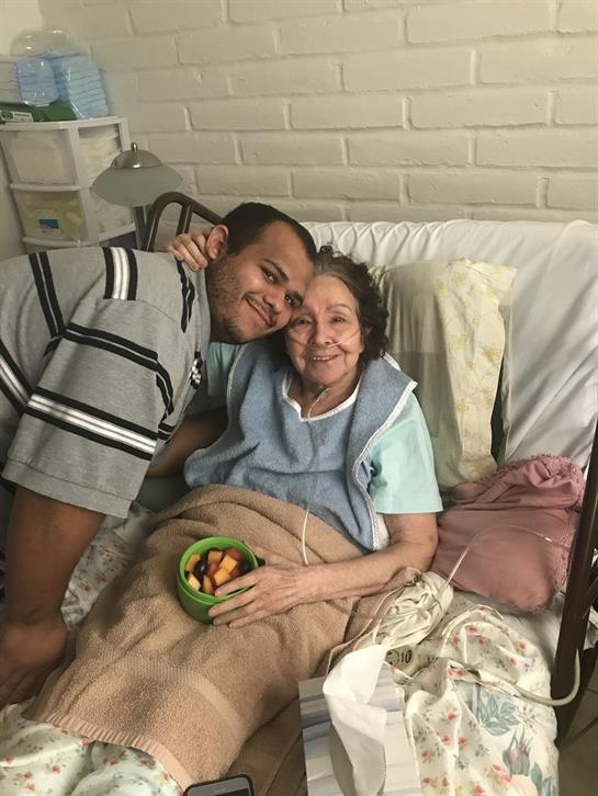 A young man shares a joyful moment with his grandmother while sitting on her bedside.