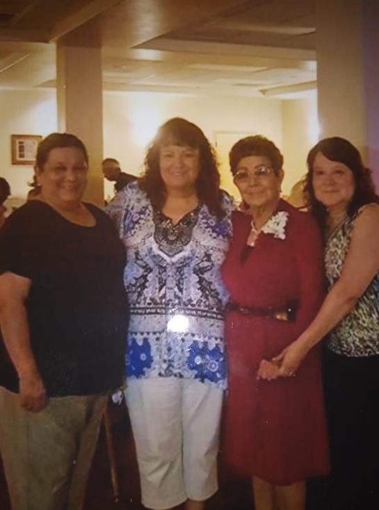Four women stand together, smiling and dressed for a celebration in a lively indoor setting.