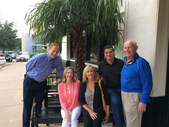 Five friends gather for a cheerful group photo outside a cafe under a palm tree in an urban area.