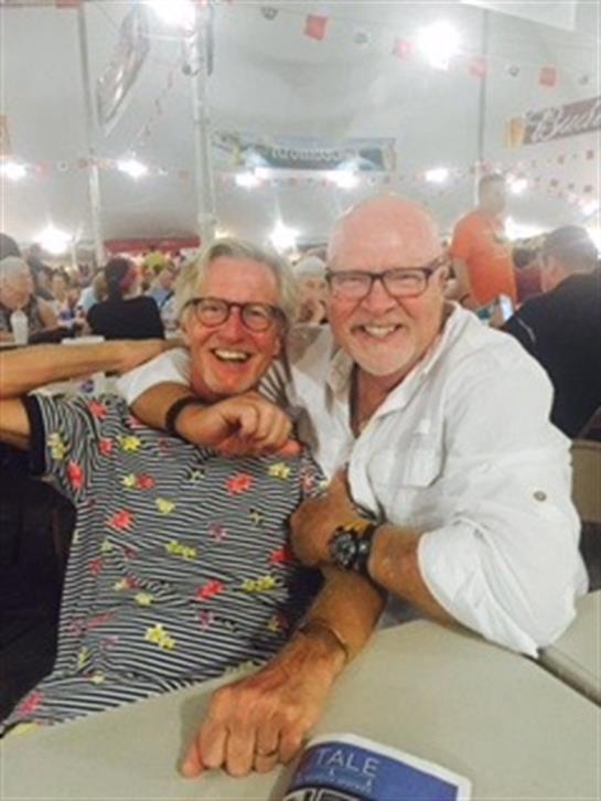 Two friends share joyful moments at a bustling festival event filled with guests.