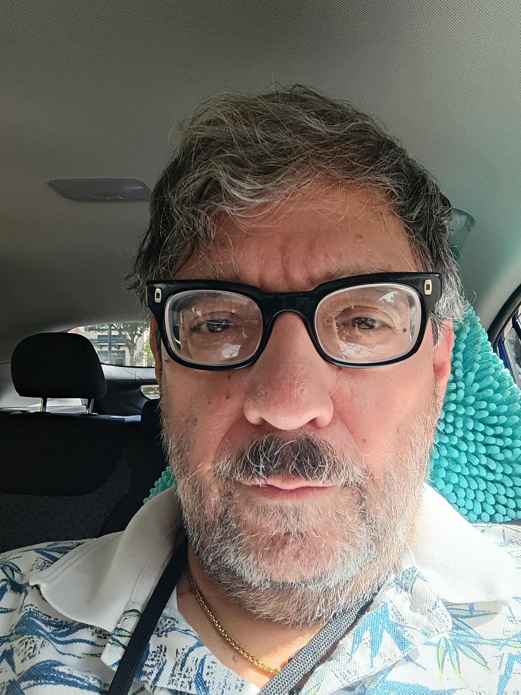 Middle-aged man with glasses takes a selfie while sitting in a car during the day.