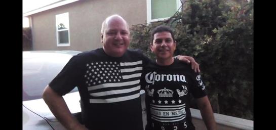 Two men outdoors smile, showcasing their shirts that reflect pride and camaraderie.