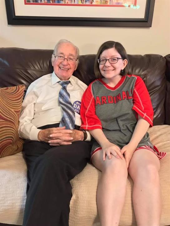 A grandfather and granddaughter smile happily while sitting closely on a couch at home.