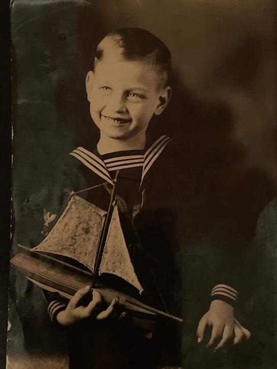 A boy dressed in a sailor outfit smiles while holding a small model sailboat.