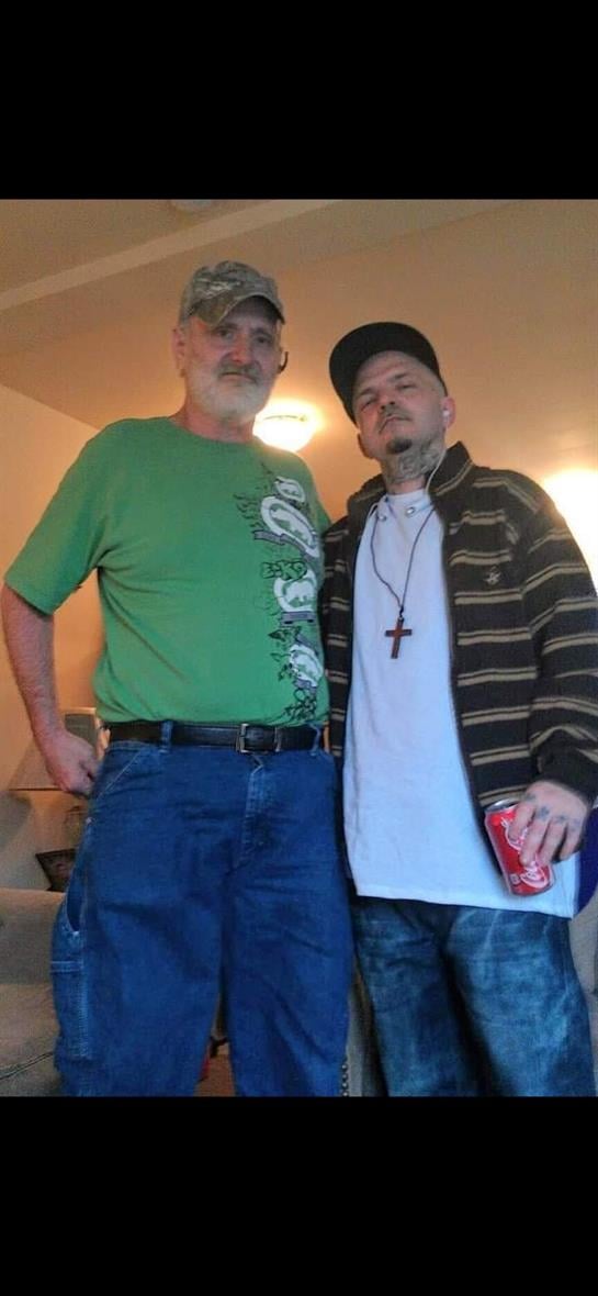 Two men stand side by side, smiling, dressed casually, enjoying their time indoors together.