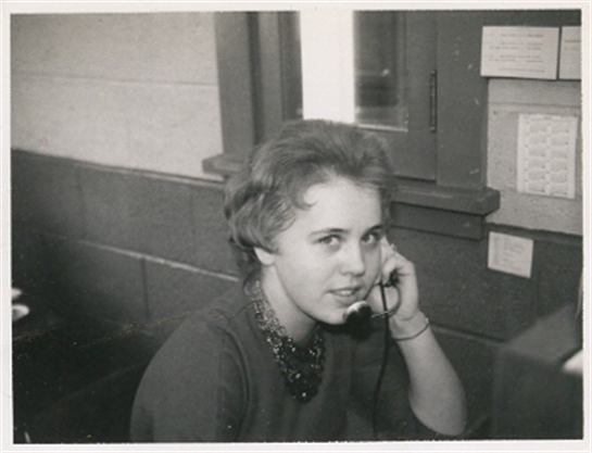 A young woman with short hair converses on a telephone in a simple office environment.