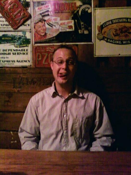 A cheerful man interacts with patrons in a rustic pub filled with vintage decor.