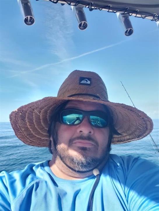 Man wearing sunglasses and a wide-brimmed hat enjoys fishing under a clear blue sky on the ocean.