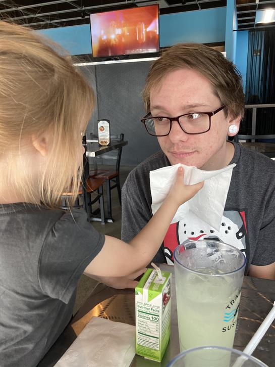Child playfully wipes the smiling adult's face as they enjoy refreshments at a table.