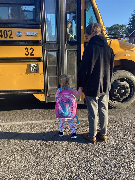 A young child in colorful attire stands with an adult near a school bus at dawn.