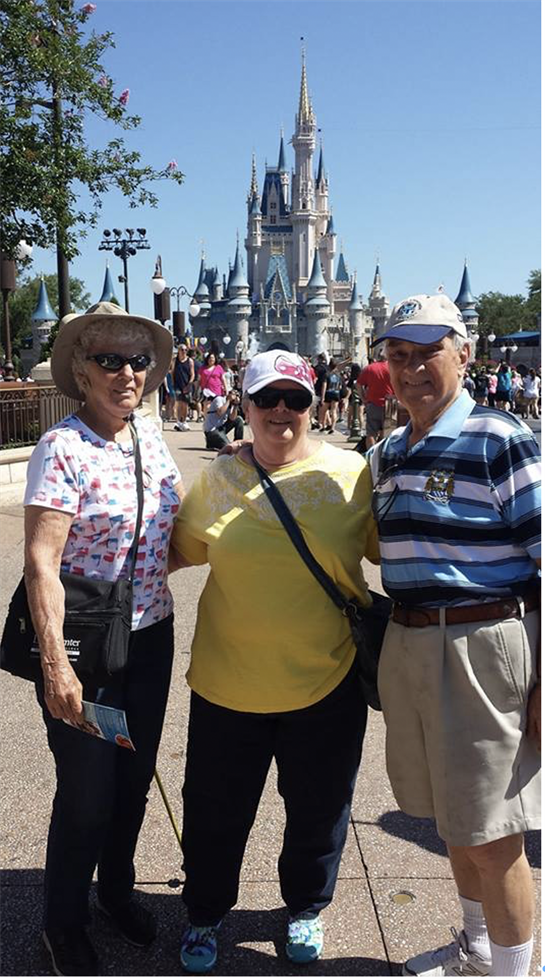 Three family members smile together in front of a grand castle on a sunny day at an amusement park.